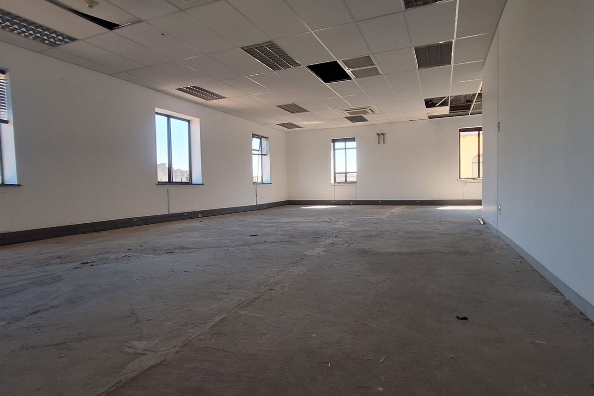 A vacant, open-plan office space featuring large windows, a suspended ceiling with missing tiles, and unfinished flooring, ready for renovation.