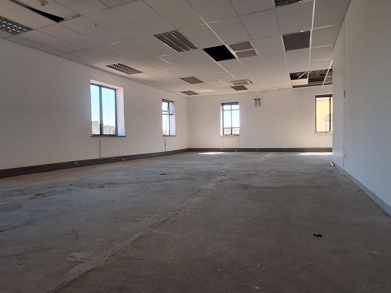 A vacant, open-plan office space featuring large windows, a suspended ceiling with missing tiles, and unfinished flooring, ready for renovation.