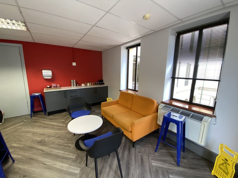 A modern office break-out area featuring a kitchenette, herringbone flooring, suspended ceiling, and large windows with venetian blinds.