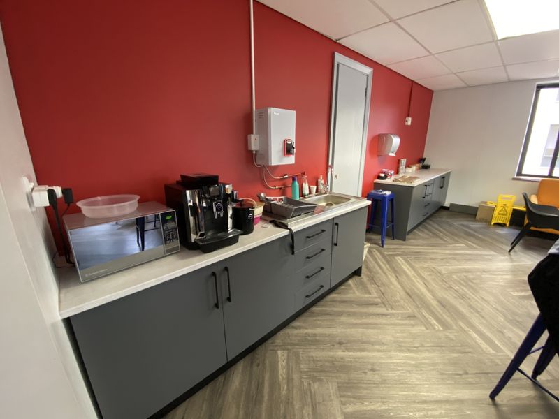 A functional office kitchenette featuring grey cabinetry, a stainless steel sink, and durable herringbone vinyl flooring against a vibrant red feature wall.
