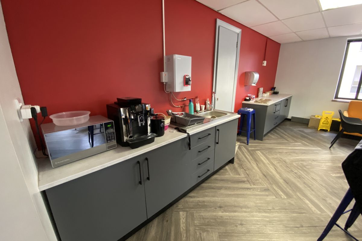 A functional office kitchenette featuring grey cabinetry, a stainless steel sink, and durable herringbone vinyl flooring against a vibrant red feature wall.
