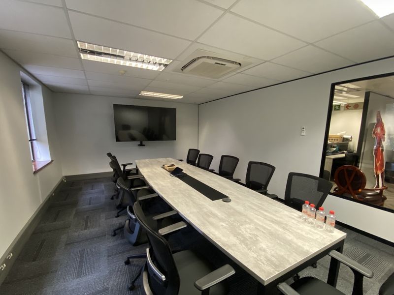 A professional boardroom or meeting room featuring a large conference table, suspended ceiling with recessed lighting, ducted air conditioning, and a glass partition wall.