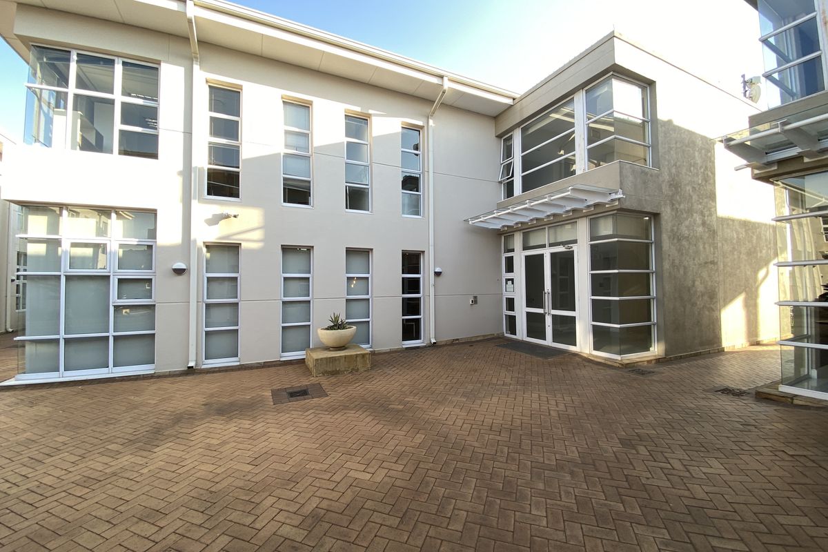 A modern office building exterior featuring a paved courtyard, large glass windows, and a double-door entrance with a protective awning.