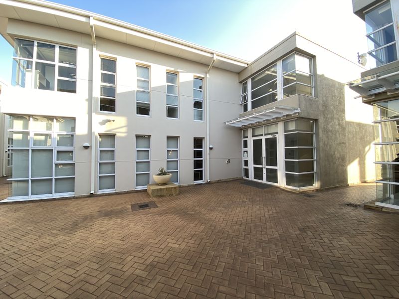A modern office building exterior featuring a paved courtyard, large glass windows, and a double-door entrance with a protective awning.