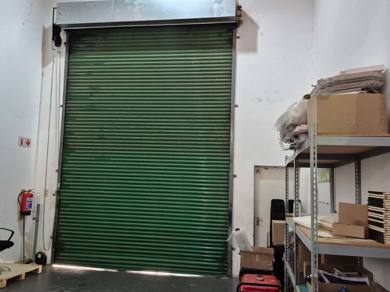 Industrial warehouse space featuring a large green roller shutter door, high ceilings, and fixed metal shelving for storage.