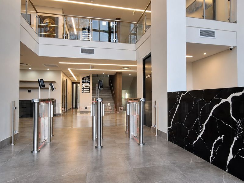 Modern office building entrance lobby featuring security turnstiles, a marble-finish reception desk, and an open-plan mezzanine level with glass balustrades.