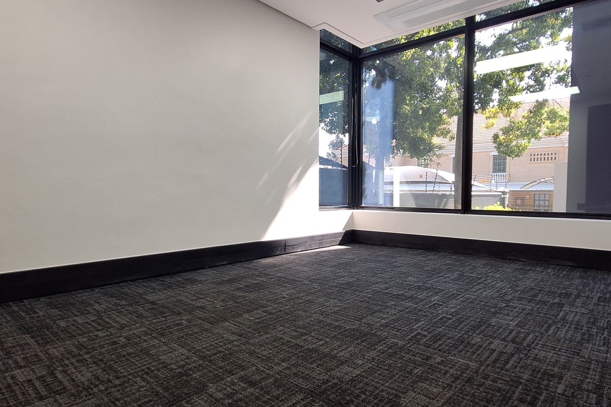 A bright, empty office space featuring large floor-to-ceiling windows, dark carpet flooring, and modern black trim finishes.