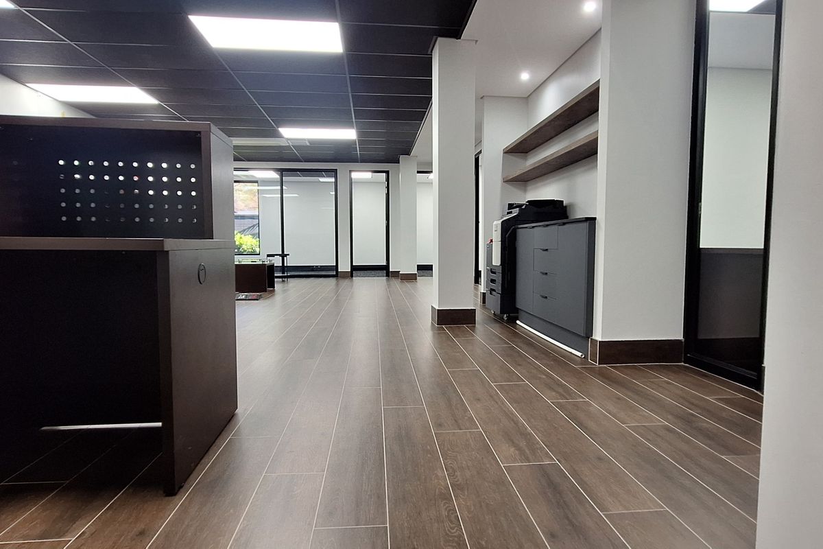 Modern open-plan office space featuring wood-look tile flooring, a suspended ceiling with recessed lighting, built-in cabinetry, and glass partition walls.