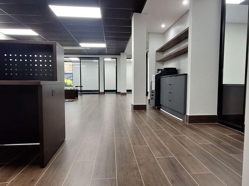 Modern open-plan office space featuring wood-look tile flooring, a suspended ceiling with recessed lighting, built-in cabinetry, and glass partition walls.