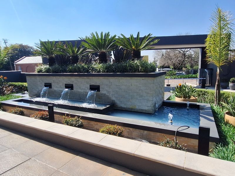 A modern office entrance featuring a sophisticated stone-clad water feature, manicured landscaping, and paved walkways, creating a professional and welcoming arrival experience.