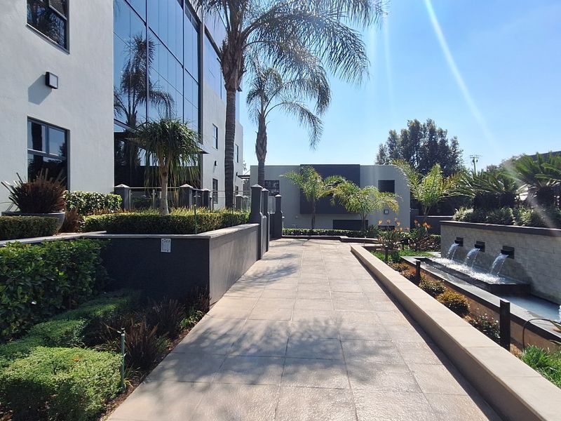 A modern office building exterior featuring a paved walkway, landscaped gardens, and a decorative water feature alongside a glass facade.