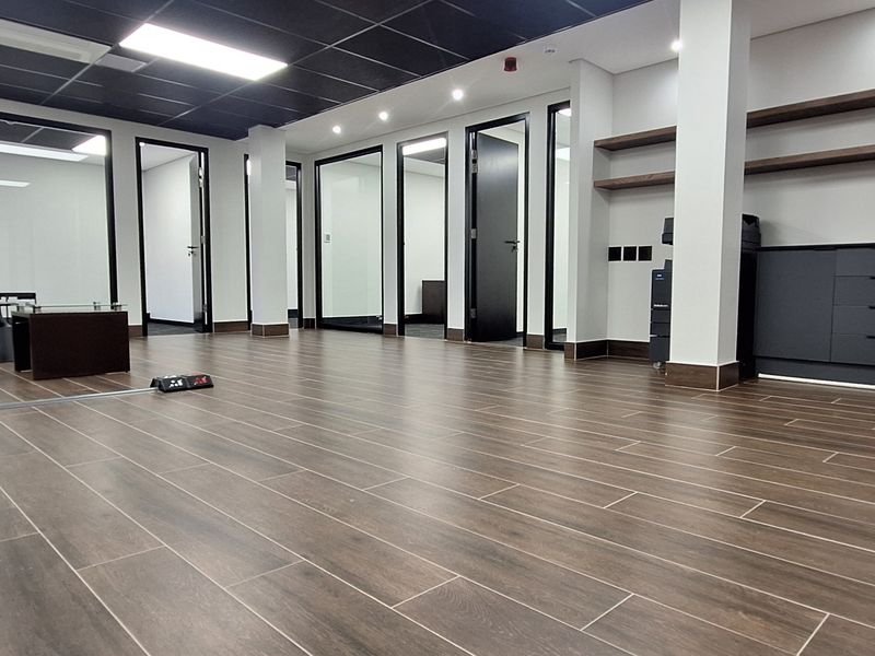 Modern open-plan office space featuring wood-look tile flooring, glass partitions, suspended ceiling with recessed lighting, and built-in storage cabinetry.