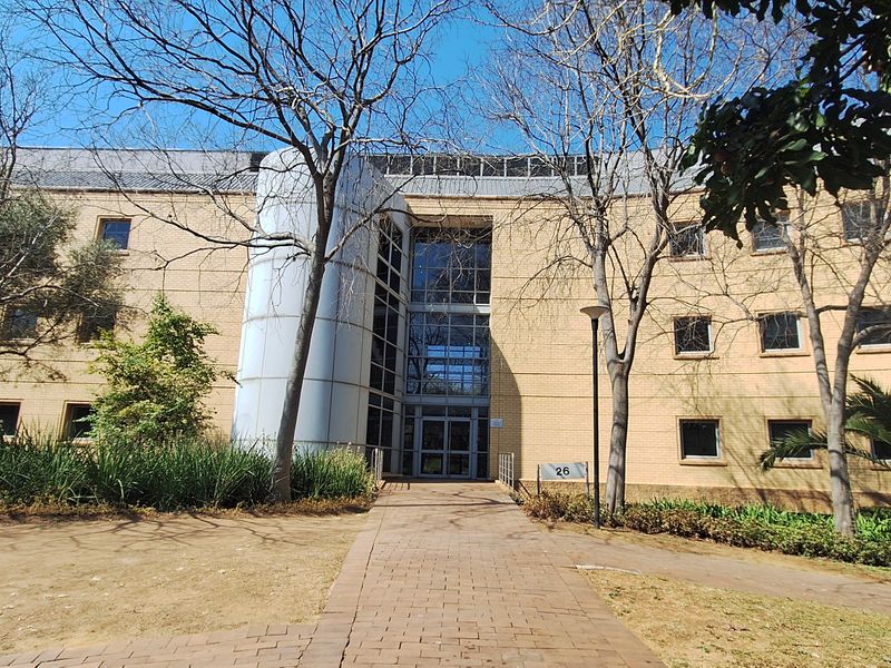 Front elevation of a multi-story office building featuring a brick facade, central glass atrium entrance, and a paved pedestrian walkway.