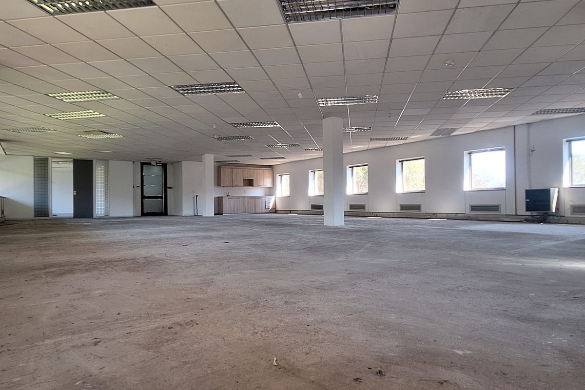 A large, open-plan office space featuring a suspended ceiling, recessed lighting, and a small built-in kitchenette area, currently awaiting flooring installation.