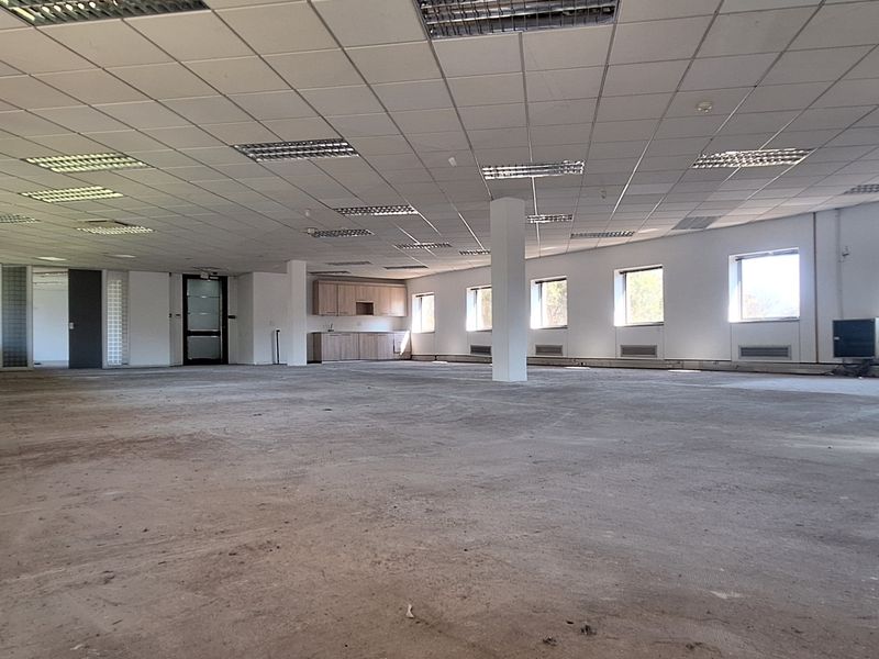 A large, open-plan office space featuring a suspended ceiling, recessed lighting, and a small built-in kitchenette area, currently awaiting flooring installation.