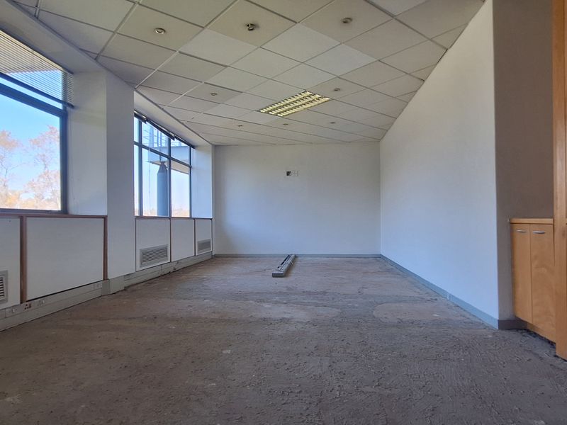 An empty office space featuring large windows, built-in wooden cabinetry, and a suspended ceiling, currently requiring flooring and ceiling repairs.