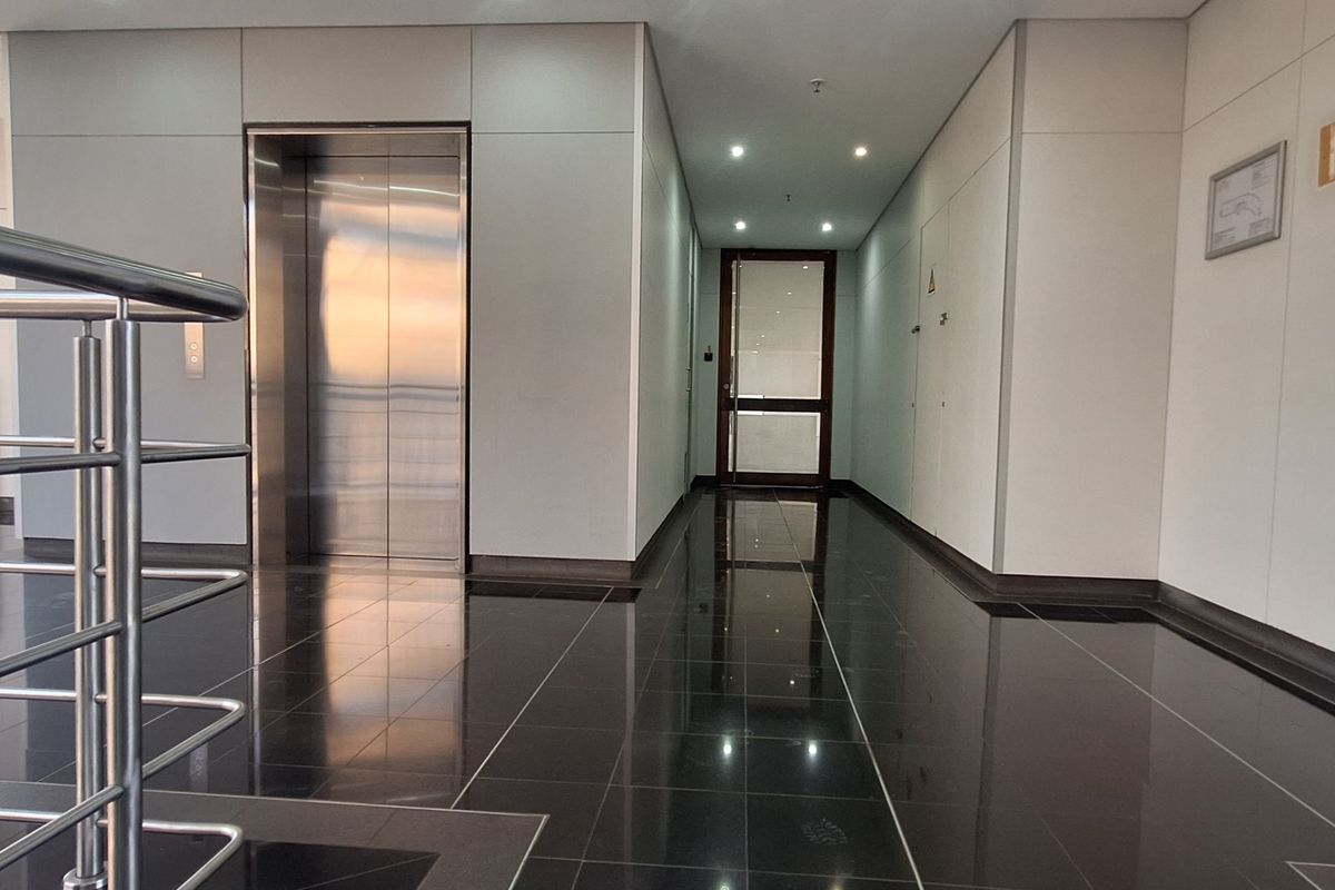 Modern office building hallway featuring polished tile flooring, a stainless steel elevator, and a sleek metal balustrade.