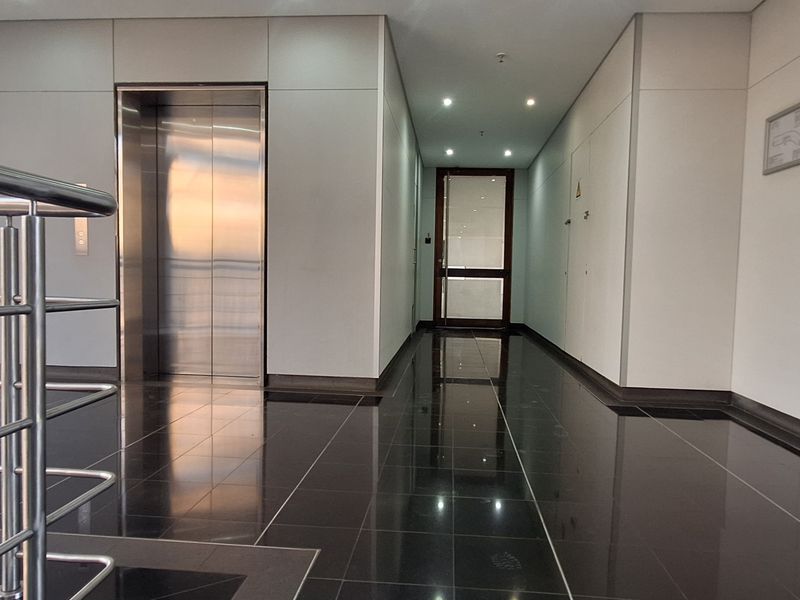 Modern office building hallway featuring polished tile flooring, a stainless steel elevator, and a sleek metal balustrade.