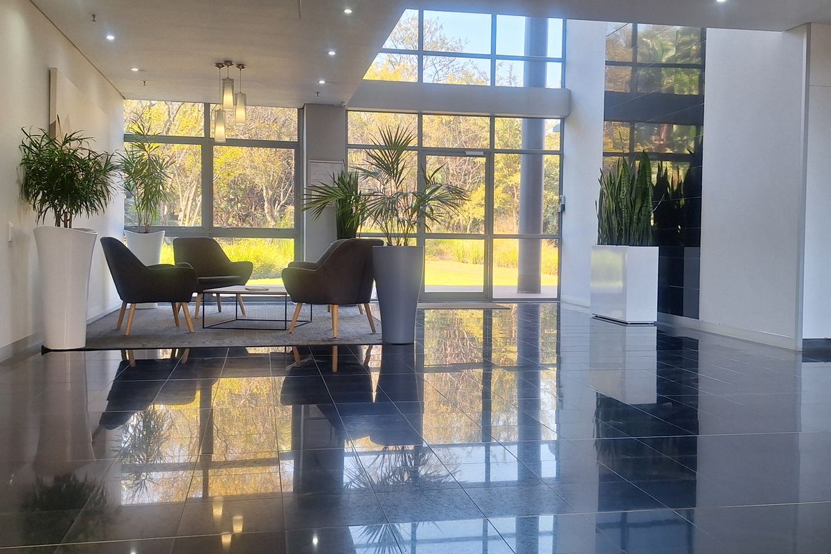 A bright and modern office building entrance lobby featuring polished dark floor tiles, expansive floor-to-ceiling windows, and a clean, professional aesthetic.