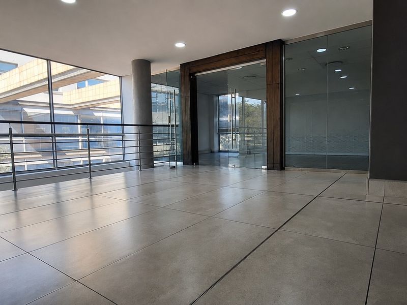 Modern office hallway featuring large format floor tiles, glass partitions, and a stainless steel balustrade, offering a bright and professional open-plan aesthetic.