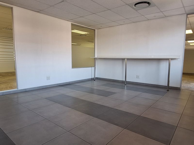 A bright, empty office space featuring ceramic floor tiles, a suspended ceiling with recessed lighting, and a built-in desk unit.