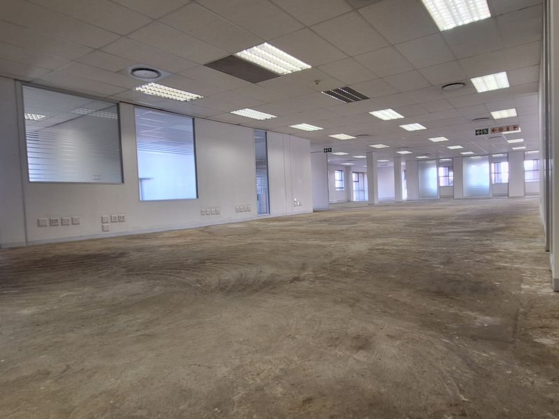 A large, open-plan office space featuring suspended ceilings, recessed lighting, and glass partitions, currently vacant with unfinished concrete flooring.