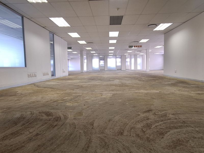An expansive, empty open-plan office space featuring glass partitions, suspended ceilings with recessed lighting, and a floor surface requiring refurbishment.