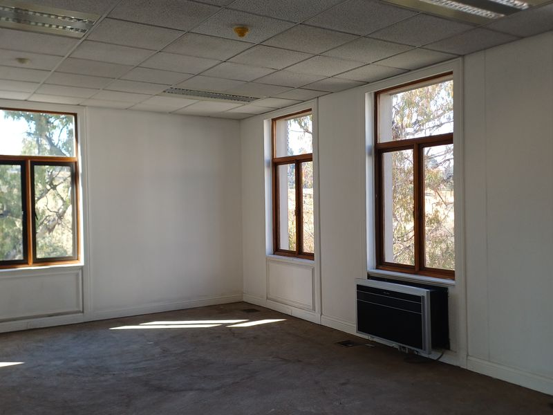 A spacious, empty office featuring large timber-framed windows, suspended ceilings with recessed lighting, and wall-mounted air conditioning units.