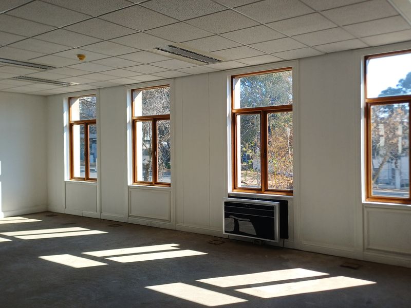 A bright, vacant office space featuring large timber-framed windows, a suspended ceiling with recessed lighting, and a wall-mounted air conditioning unit.