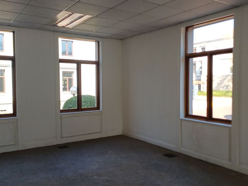 An empty, well-lit office space featuring large timber-framed windows, a suspended ceiling with recessed lighting, and carpeted flooring.