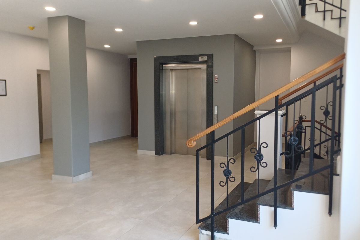 Modern office building lobby featuring a stainless steel elevator, tiled flooring, recessed lighting, and a staircase with a decorative wrought iron balustrade.