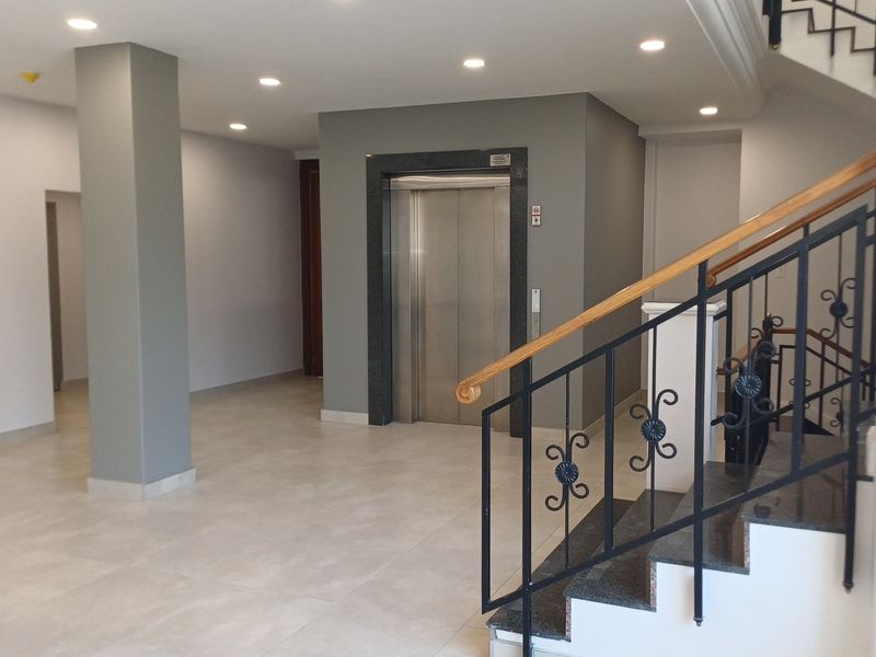 Modern office building lobby featuring a stainless steel elevator, tiled flooring, recessed lighting, and a staircase with a decorative wrought iron balustrade.