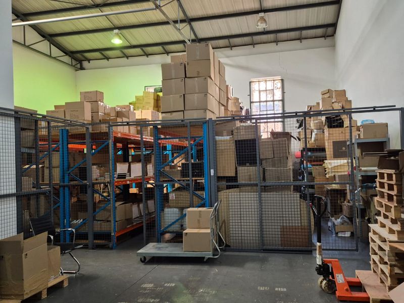 Industrial warehouse space featuring high ceilings with exposed steel trusses, concrete flooring, and secure metal mesh partitioned storage areas.