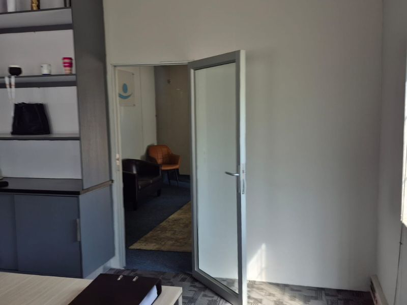 An office space featuring built-in shelving, carpeted flooring, and an aluminium-framed door leading to an adjacent room with security-barred windows.