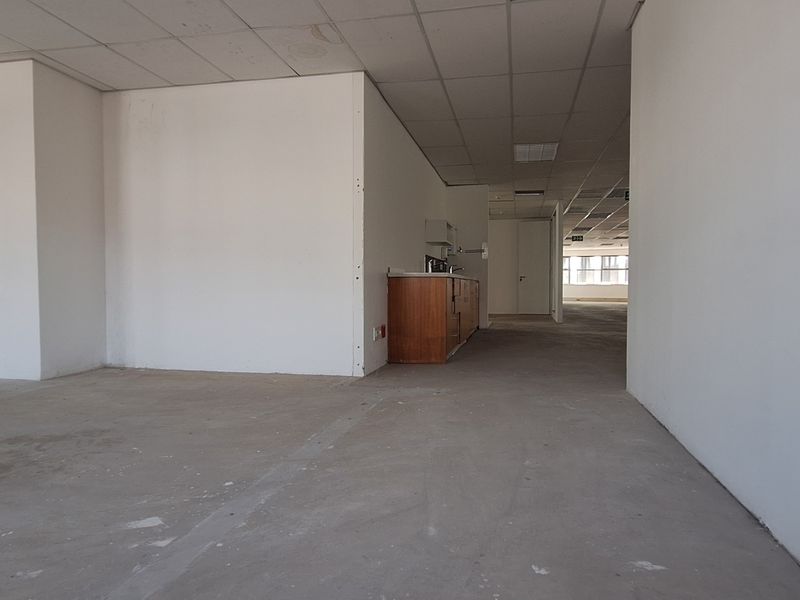 An open-plan office space featuring a suspended ceiling, recessed lighting, and a small built-in kitchenette area with unfinished concrete flooring.