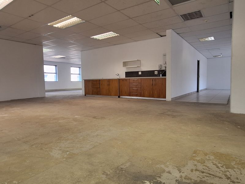 Open-plan office space featuring a kitchenette, glass partitions, and suspended ceilings, currently vacant with worn concrete flooring.
