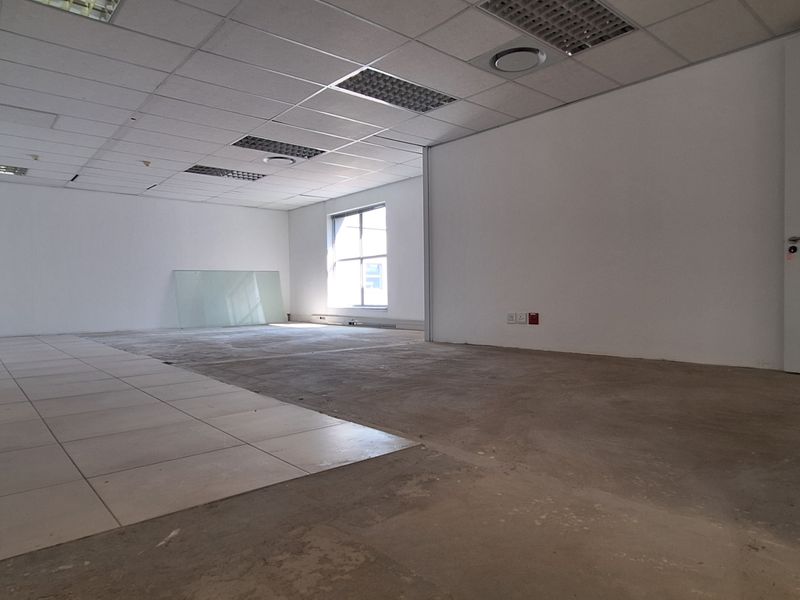 An empty, open-plan office space featuring a suspended ceiling, recessed lighting, and a mix of tiled and unfinished concrete flooring.