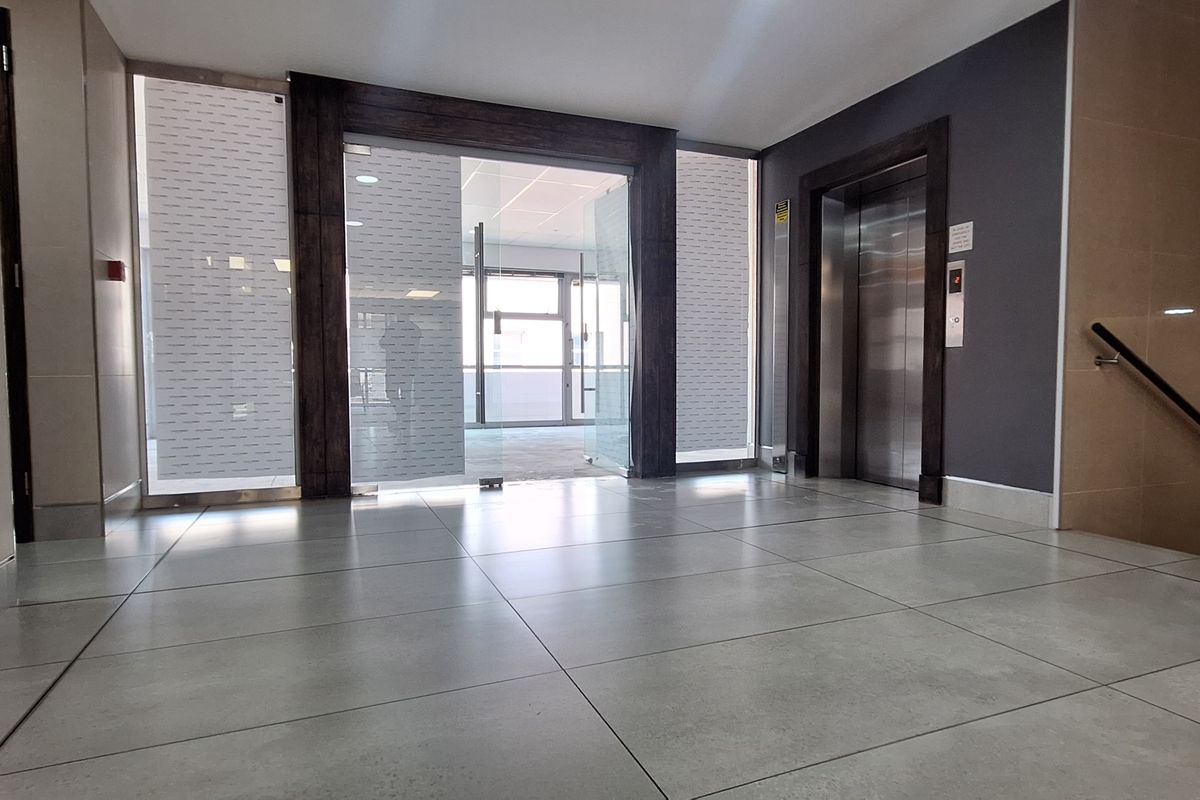 Modern office building entrance lobby featuring tiled flooring, a stainless steel elevator, and glass entry doors leading to the main office space.