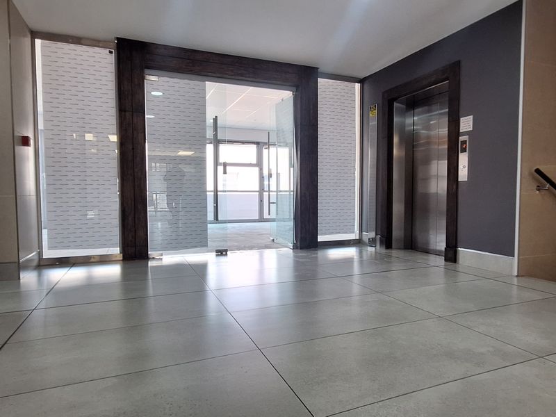 Modern office building entrance lobby featuring tiled flooring, a stainless steel elevator, and glass entry doors leading to the main office space.