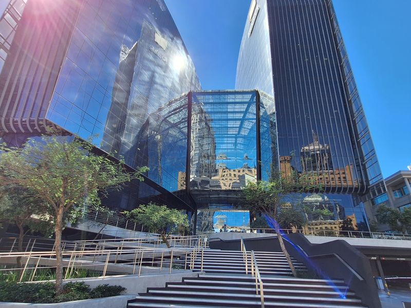 Modern commercial office building featuring a striking glass facade, central atrium entrance, and tiered paved pedestrian access with integrated steel handrails.