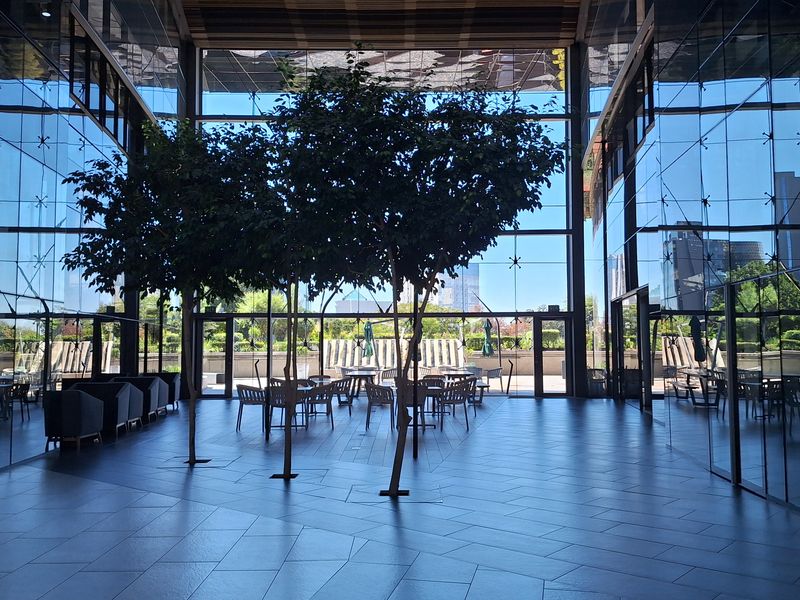 Modern open-plan office atrium featuring floor-to-ceiling glass walls, high ceilings with recessed lighting, and sleek tiled flooring with integrated indoor trees.