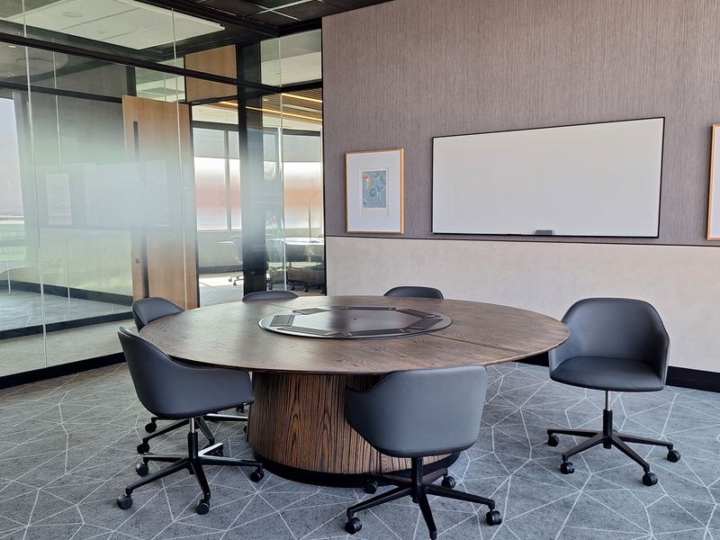 A modern, professional office meeting room featuring glass partitions, a circular conference table, and contemporary carpet flooring.