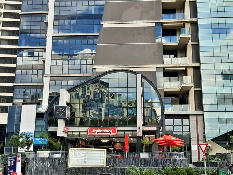 Modern multi-story commercial office building featuring a prominent glass facade, integrated balconies, and ground-level retail space with a paved terrace area.