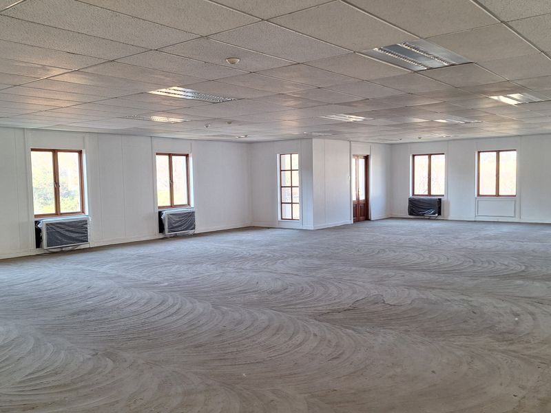 A large, open-plan office space featuring a suspended ceiling, recessed lighting, and multiple windows with integrated air conditioning units.