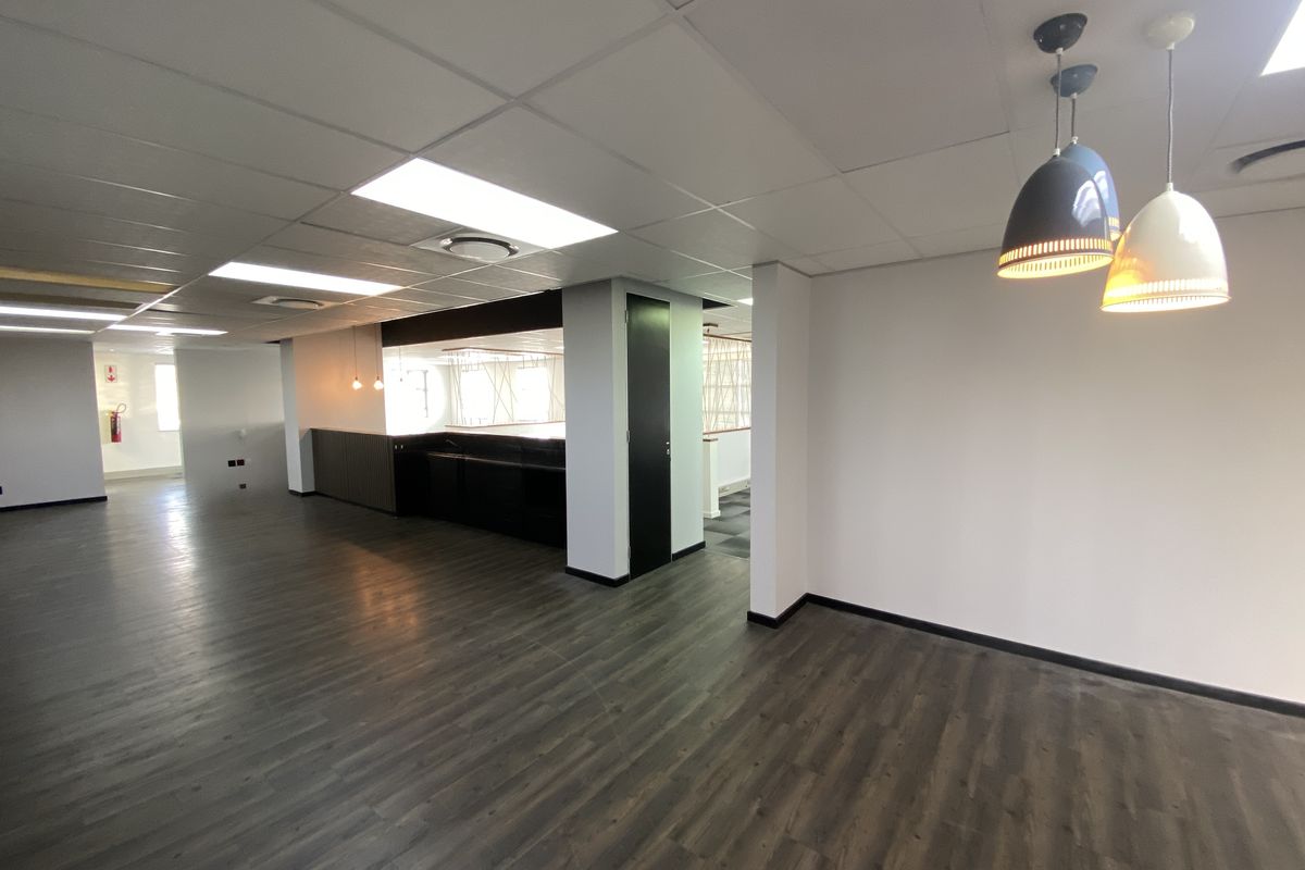 A spacious, modern open-plan office area featuring dark laminate flooring, a suspended ceiling with recessed lighting, and a sleek built-in kitchenette.