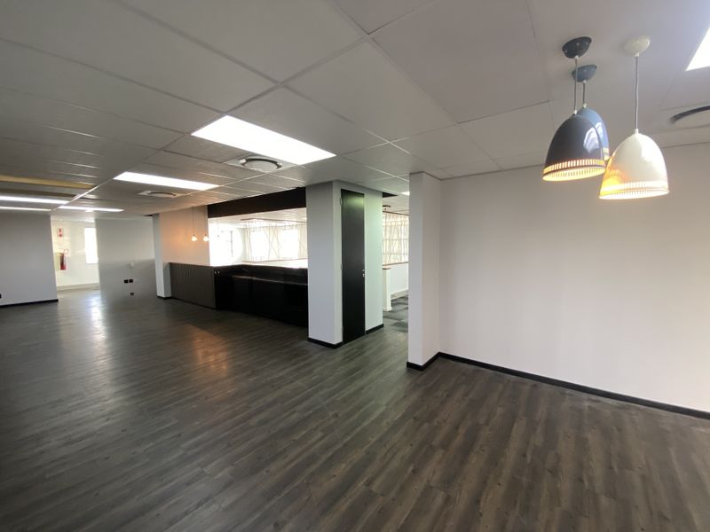 A spacious, modern open-plan office area featuring dark laminate flooring, a suspended ceiling with recessed lighting, and a sleek built-in kitchenette.
