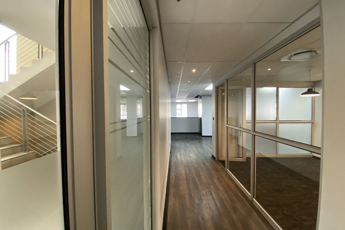 A modern office hallway featuring laminate flooring, suspended ceilings with recessed lighting, and glass partition walls leading to adjacent office spaces.