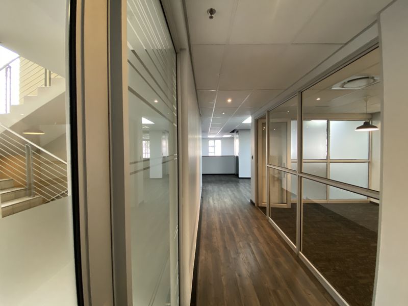 A modern office hallway featuring laminate flooring, suspended ceilings with recessed lighting, and glass partition walls leading to adjacent office spaces.
