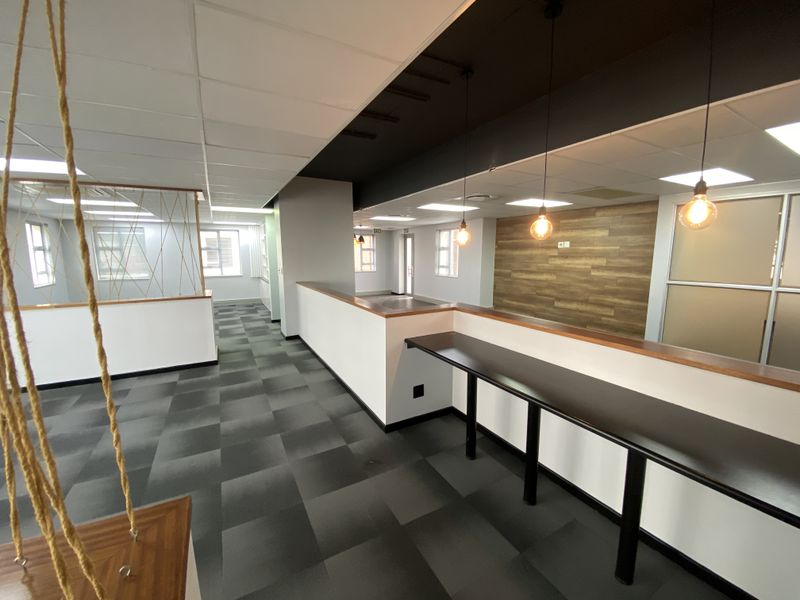 A modern, open-plan office space featuring carpet tiles, a built-in desk, wood-paneled accent wall, and contemporary pendant lighting.