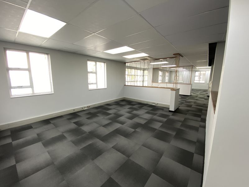 Spacious open-plan office area featuring neutral walls, carpet tiles, suspended ceilings with recessed lighting, and large windows providing ample natural light.